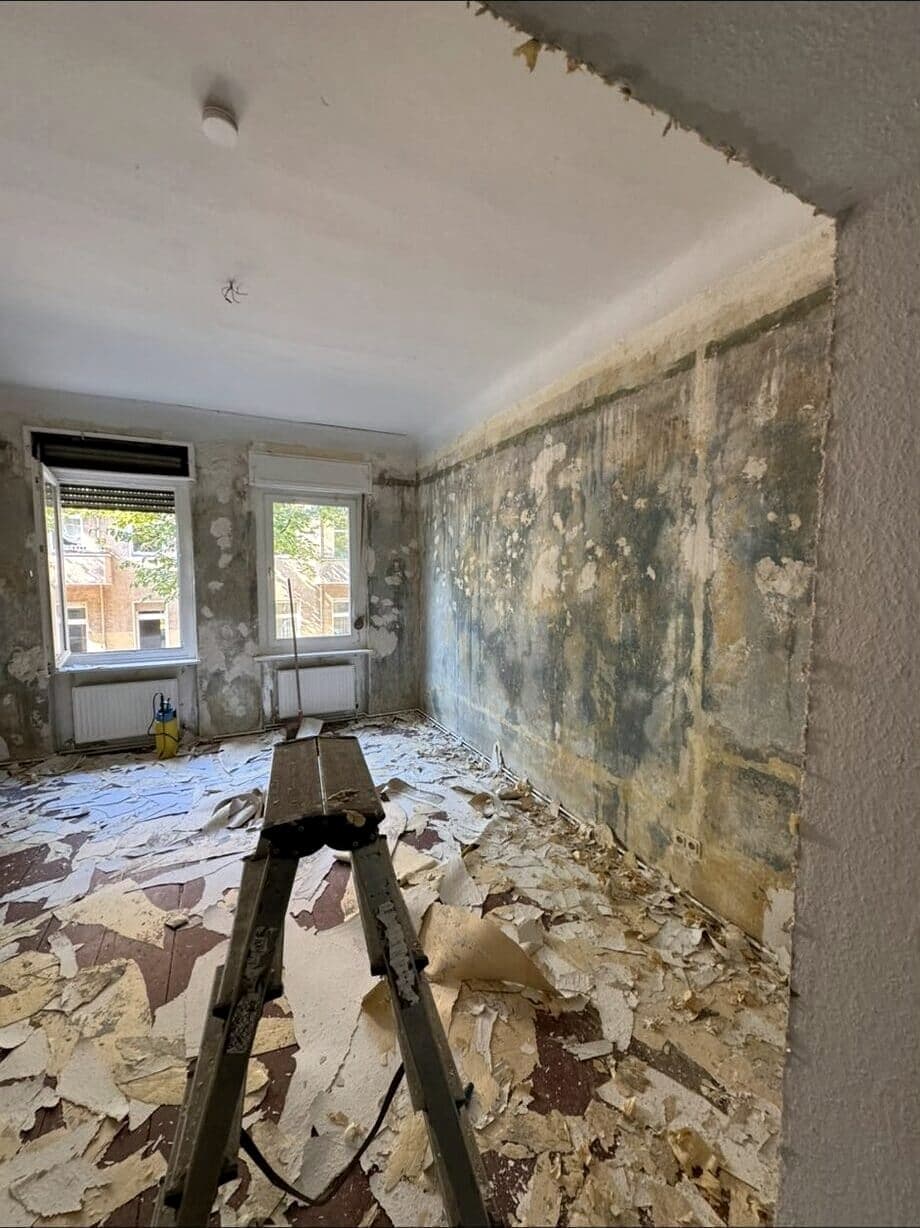 Wohnraum vor der Sanierung: abgelöste Tapeten, fleckiger Altputz und Bauschutt auf dem Boden